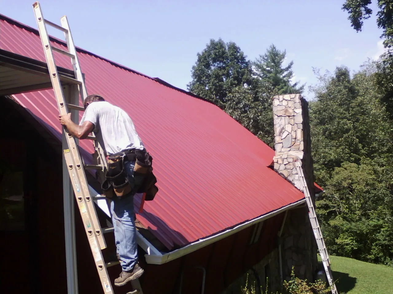 Roofer on a ladder beside a red metal roof and stone chimney — Soffit & Fascia Repair by Roofing Brothers in Morris